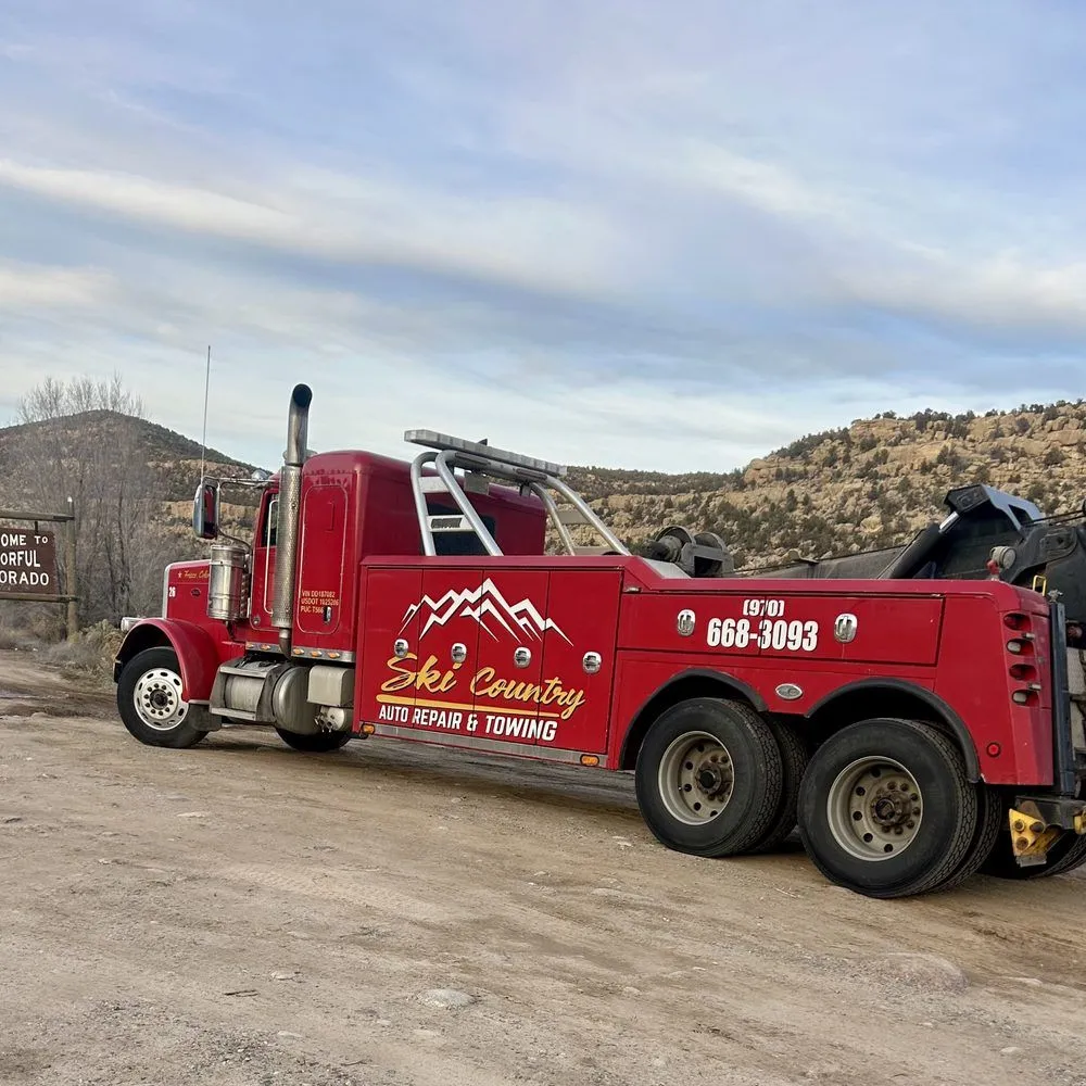 Red tow truck parked on a dirt road near mountains under a cloudy sky.