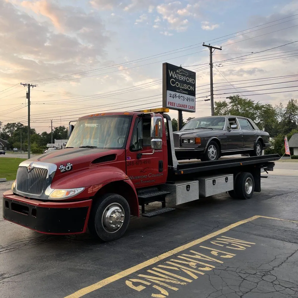 Red flatbed tow truck hauling a dark classic sedan, parked on a street at dusk