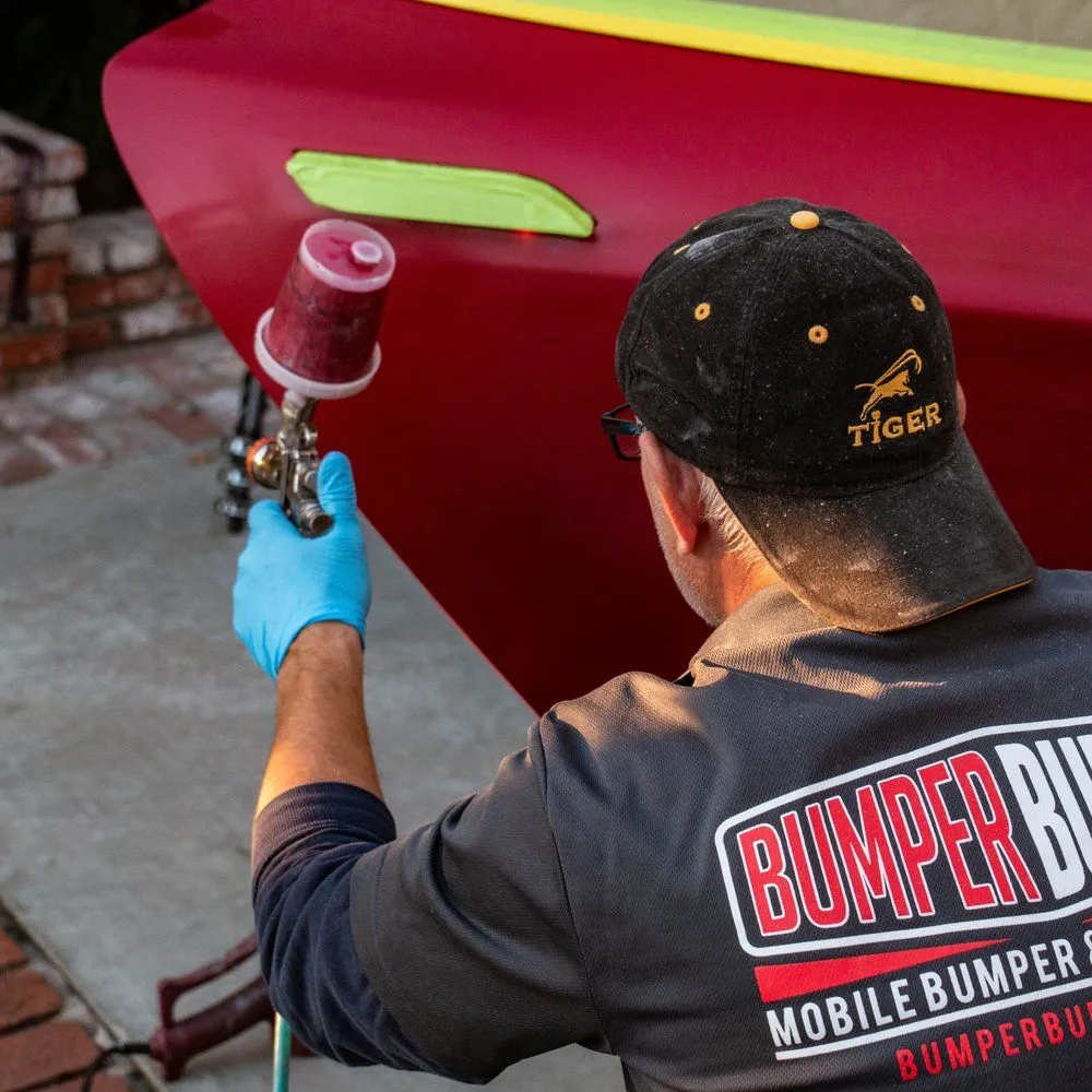 Technician in blue gloves spray-painting a red vehicle bumper with a handheld paint gun