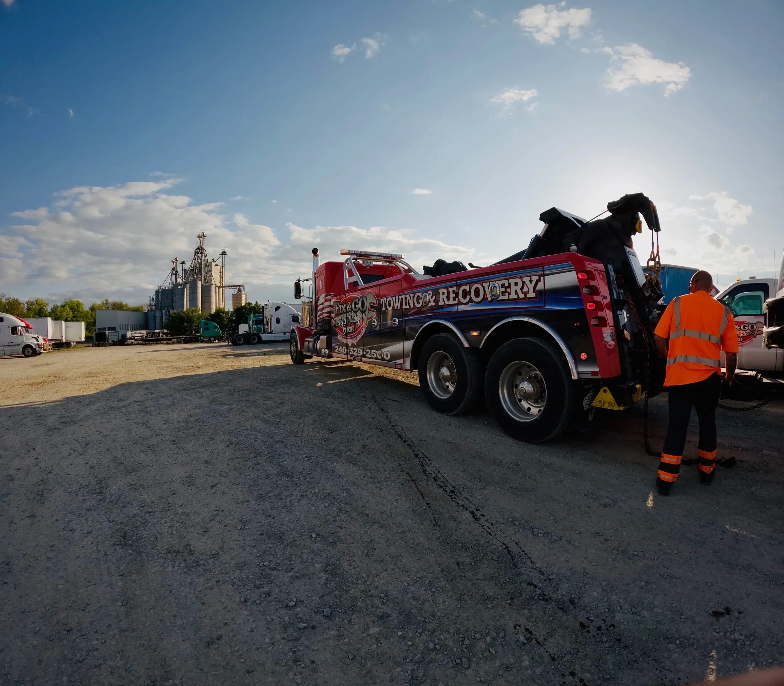 Tow operator in orange hi-vis gear beside a red heavy wrecker on a gravel lot